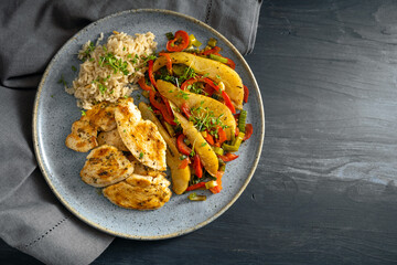 Fried chicken slices with whole grain rice and vegetables of bell pepper, pear and leek on a blue plate and a dark gray wooden table, copy space, top view from above