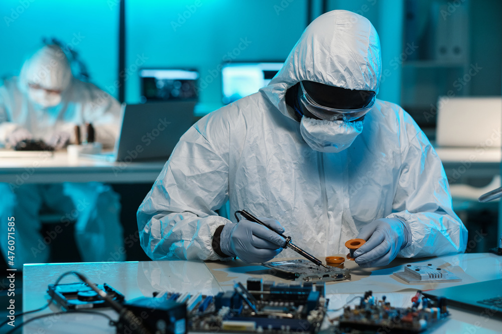 Poster contemporary technician in protective workwear soldering tiny parts of circuit board while sitting b