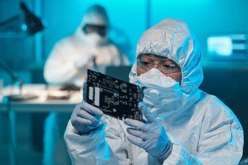 Young Asian female in protective workwear looking at circuit board in her gloved hands while...