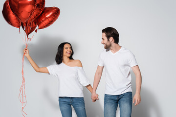 happy young couple in white t-shirts holding hands and looking at each other near heart-shaped...