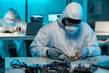 Contemporary technician in protective workwear soldering tiny parts of circuit board while sitting...