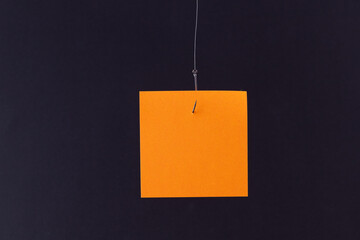 Mockup of a Blank Orange Memo Paper with Copy Space Hanging on a Fishing Hook Against the Black Background. Sticky Note Template
