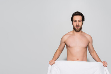 Fototapeta premium astonished shirtless man holding white blanket while looking at camera on grey.
