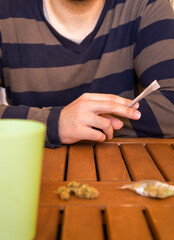 Unrecognized man with a cannabis joint sitting at table having a drink at a party at an outdoor location.