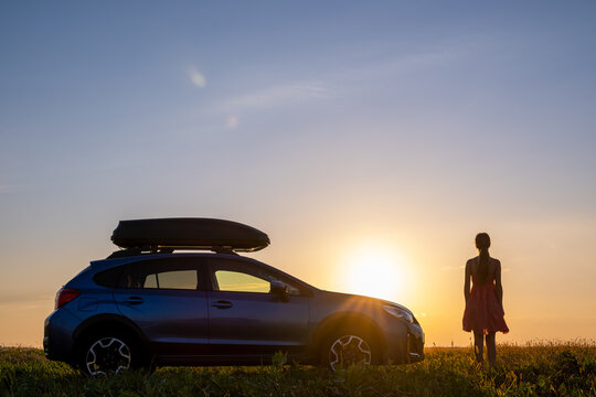 Dark silhouette of lonely woman relaxing near her car on grassy meadow enjoying view of colorful sunrise. Young female driver resting during road trip beside SUV vehicle.