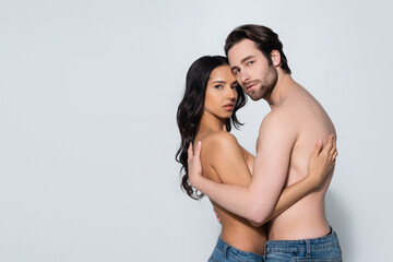 sensual couple in jeans only embracing and looking at camera on grey.