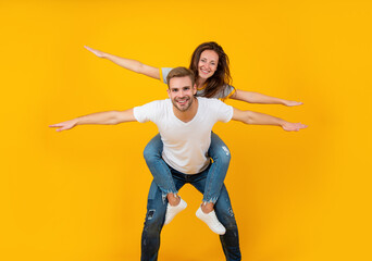 Happy playful woman and man pretend flying doing piggyback ride yellow background, fun