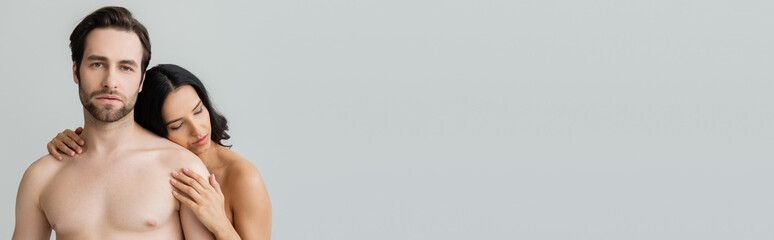 Fototapeta premium young shirtless man looking at camera near brunette woman embracing him isolated on grey, banner.