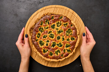 Women's hands hold quiche pie with broccoli, tuna and cheese on round wooden cutting board on black...