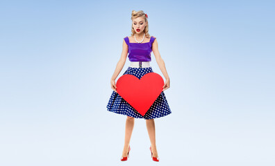 Full body of woman holding heart symbol, dressed in pin-up style dress with polka dot, over light blue background. Blonde pinup girl posing in retro fashion and vintage concept studio shot.