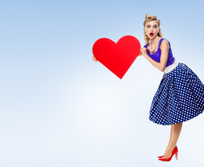 Full body of woman holding heart symbol, dressed in pin-up style dress with polka dot, over light blue background. Blonde pinup girl posing in retro fashion and vintage concept studio shot.
