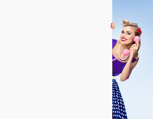 woman with phone, in pin-up style dress, showing blank signboard with copyspace area, over light blue background. Blonde pinup girl posing in retro fashion and vintage concept studio shot.