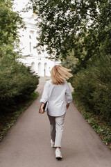 Fototapeta na wymiar blonde in white clothes walks through the park to the university with a book in her hands green plants trees white building. vertical