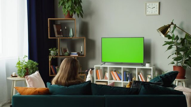 Woman watching television with chroma green screen. Unrecognizable female person switching channels on tv with remote controller, sitting on sofa in living room. Spare time at home, relaxing.