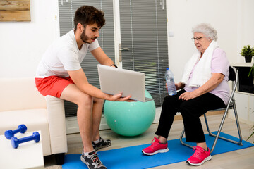 senior elderly woman with a personal trainer private fitness coach at home handsome young man