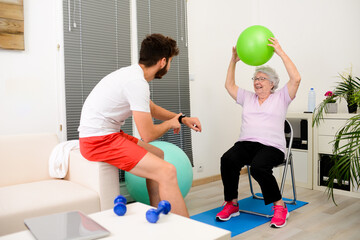 senior elderly woman with a personal trainer private fitness coach at home handsome young man