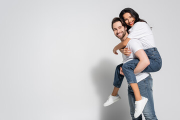 cheerful young couple in white t-shirts and jeans looking at camera while having fun on grey.