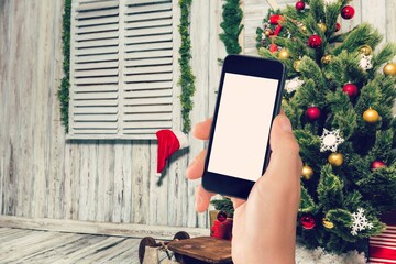Hand of human holding the phone with empty screen mockup near decorated festive tree, celebrating...