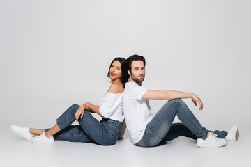 full length view of young couple in white t-shirts and jeans sitting back to back on grey.