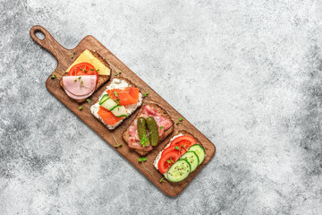 Various sandwich on a wooden board, gray grunge background. Open sandwiches with vegetables, bacon, salmon and ham. Different bruschetta. Top view, flat lay.