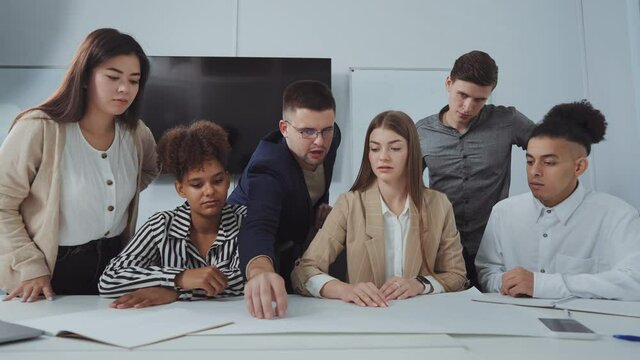 Young successful team of designers discussing plan on paper lying on table, men and women expressing ideas and showing at certain spots on plan. Concept of business