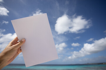 Woman's hand holding blank white paper in front of sea and blue sky background. Mockup.