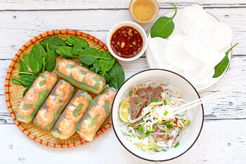 Close-up of Vietnamese spring rolls with chicken, tiger prawns, Pho bo, Vietnamese soup with noodles and beef, fresh herbs, rice chips on a wooden board. Sesame sauce. A Variety Of Vietnamese Food.