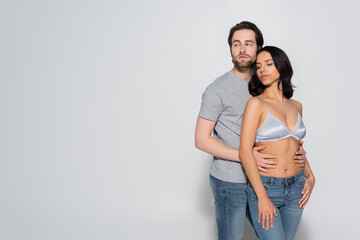 Fototapeta premium young man in t-shirt looking away while hugging hot woman in satin bra on grey.
