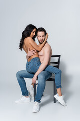 shirtless man in jeans sitting on chair with passionate girlfriend on grey.