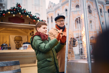 Little boy and his grandfather warm their hands by the fire at Christmas market.