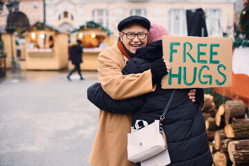 Happy senior man offers free hugs while holding cardboard sign and embracing a stranger at Christmas market.
