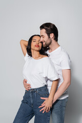 Naklejka premium smiling man hugging seductive woman standing with hand behind head and closed eyes on grey.