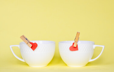 two little red paper heart attached by the edge of a ceramic white cup with wooden decorative clothes pin. valentines day, i love. love and drink coffee, tea, cappuccino. yellow background. february. 