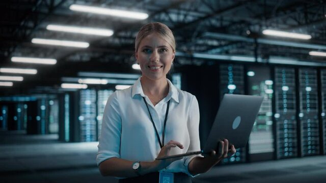 Portrait of Successful Female Chief Technology Officer Using Laptop Computer, Standing in Big Warehouse Data Center, Looking at Camera. System Administrator Maintenance of SAAS, Cloud Services Server