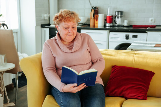 Overweight Woman Reading A Book Sitting On The Couch In The Living Room.