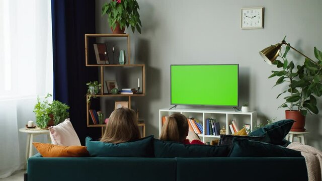 Women watching television with chroma green screen. Girlfriends switching channels on tv with remote controller, sitting on sofa together in living room. Spare time at home, relaxing.