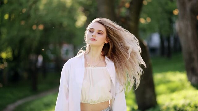 Beautiful Sexy Young Blonde Woman with Clean and pefect Skin relaxing in the park. Portrait of university student looking at camera. Woman Standing on the Wind. Breeze is playing with girls hair