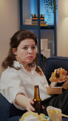 Caucasian person relaxing on sofa while eating hamburger and drinking beverage at home. Woman with fast food delivery meal watching movie on television and having unhealthy lifestyle
