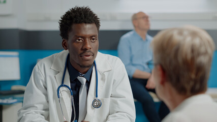 Close up of specialist sitting at desk wearing white coat while talking to elderly patient in healthcare office. Doctor with stethoscope and laptop discussing illness to treat senior woman