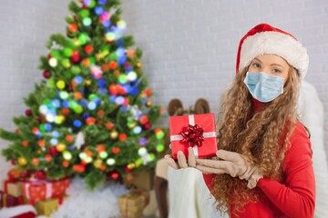 Portrait of a beautiful girl in medical mask and santa hat with a present in hands, standing in front of decorated christmas tree at home