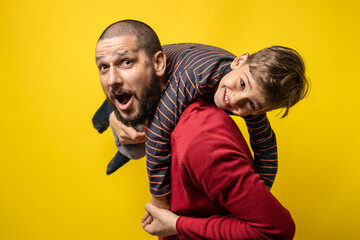 Fototapeta premium Adult caucasian man with beard holding his small son four years old boy on his shoulder while posing in front of yellow background real people family parenthood and growing up concept