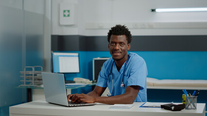 Portrait of male nurse with uniform and stethoscope using laptop on desk sitting in medical cabinet...