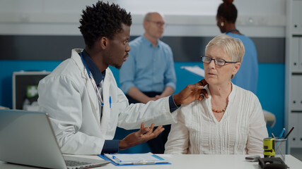 Fototapeta premium Young medic consulting old person for checkup appointment sitting at desk in healthcare cabinet. Doctor advising senior woman about treatment while nurse and elder man talking in background