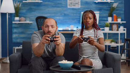 Modern interracial couple playing video game on TV with console and joysticks at home. Multi ethnic people having fun with controller and technology in living room, looking at camera