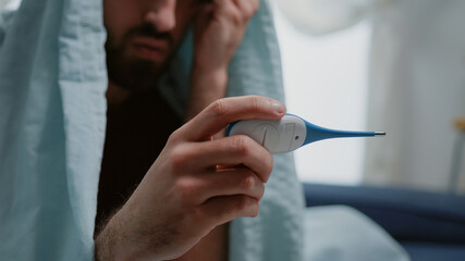 Close up of sick man looking at thermometer measuring fever and flu symptoms. Adult with virus infection using medical device to check temperature wrapped in blanket, feeling cold.