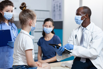 Fototapeta premium African american pediatrician doctor with protection face mask against coronavirus explaining medical treatment to family during clinical appointment in hospital office. Health care service