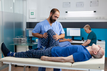 Physiotherapist helping old man with leg injury to recover at kinetotherapy clinic. Chiropractor giving support to retired patient with knee pain and muscle pressure for recovery.