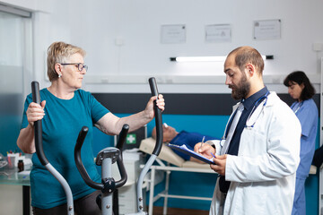 Old patient doing physical activity with stationary bicycle while doctor giving assistance for physiotherapy. Retired woman using bike for fitness workout to recover from injury.