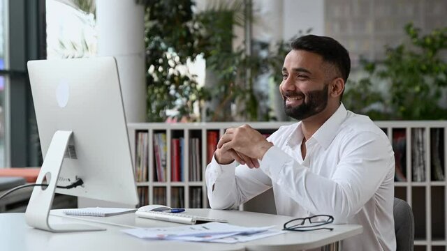 Happy friendly, confident successful Indian businessman or CEO with a beard, sitting in an office, wearing a white shirt, using a computer for personal communication, waving his hand, smiling