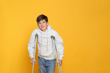 Teenage boy with injured leg using crutches on yellow background, space for text